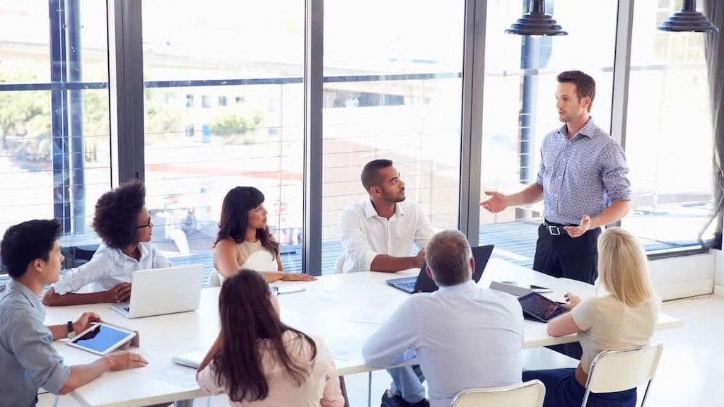 A business team engaged in discussion around a meeting table, with a presenter standing.