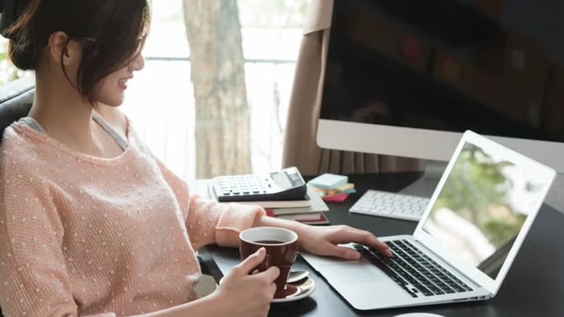 A woman works from home on her laptop with a coffee in hand, surrounded by office essentials on her desk.
