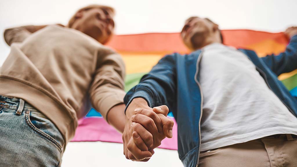 A couple holding hands beneath a rainbow flag, symbolizing LGBTQ+ pride and solidarity.