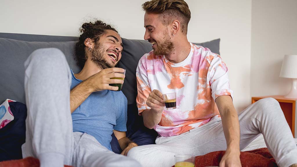 A couple sitting together on a bed, laughing and enjoying drinks, capturing a joyful and intimate moment.