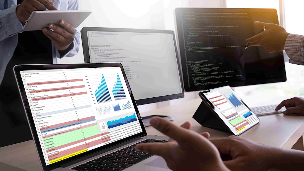 A professional works on a laptop, analyzing reports and data, with multiple screens showing graphs and metrics.