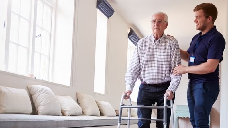 A caregiver assisting an elderly man with a walker inside a bright, modern room.