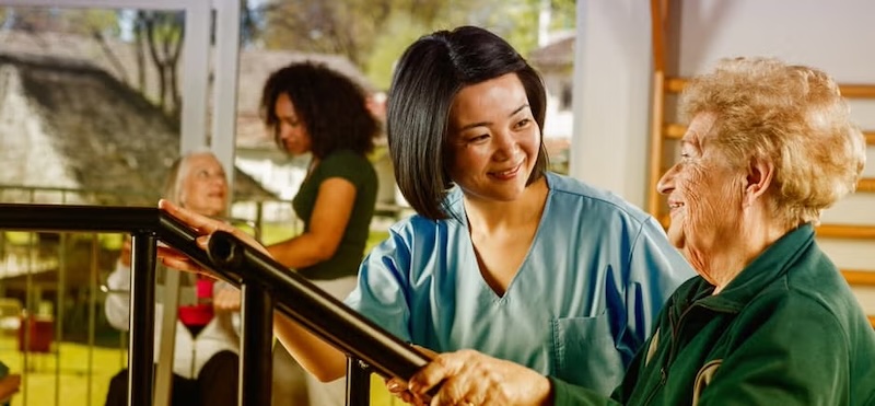 A caregiver assisting an elderly woman during physical therapy, with other residents and staff in the background.