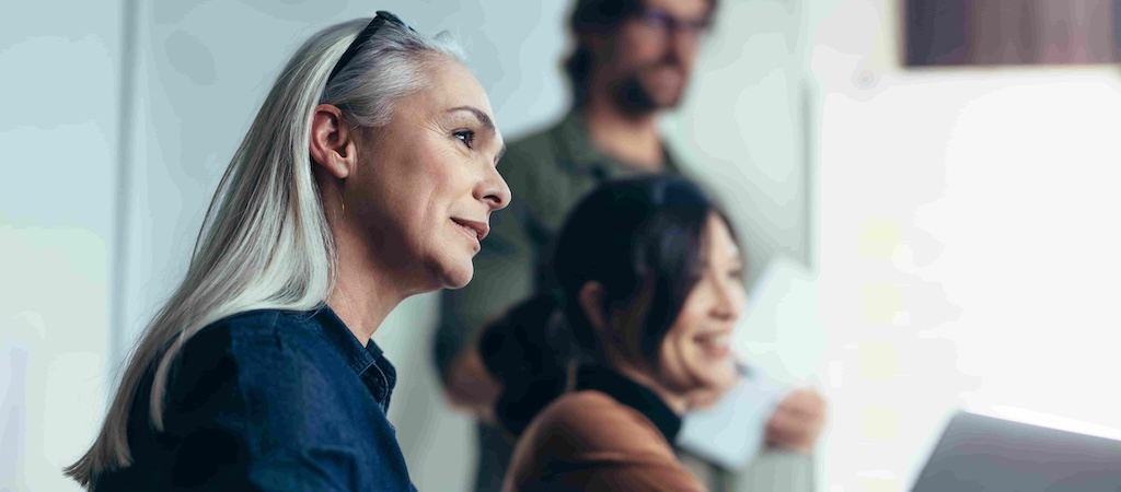 A group of professionals engaged in a meeting, with a senior woman and another person smiling attentively.