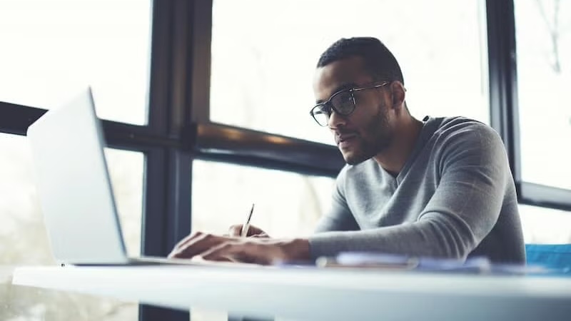 A man in glasses working intently on his laptop at a sunlit desk.