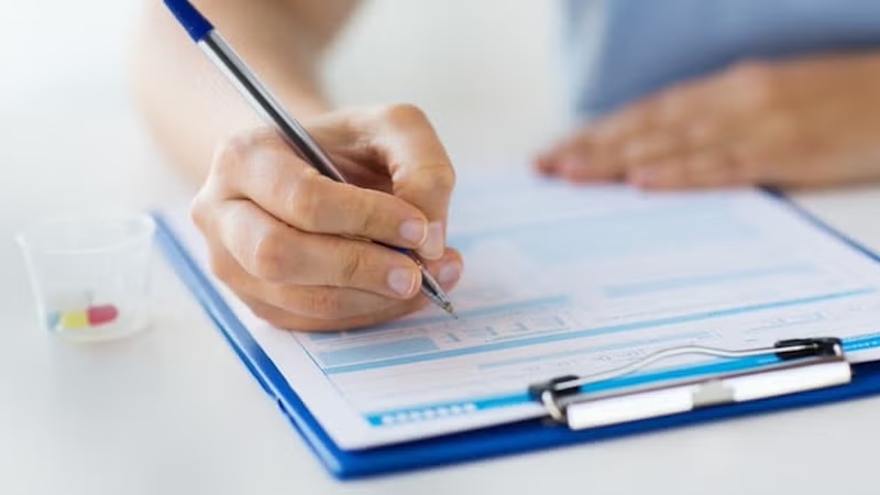 A person filling out a form on a clipboard with a pen, with a small medicine cup nearby.