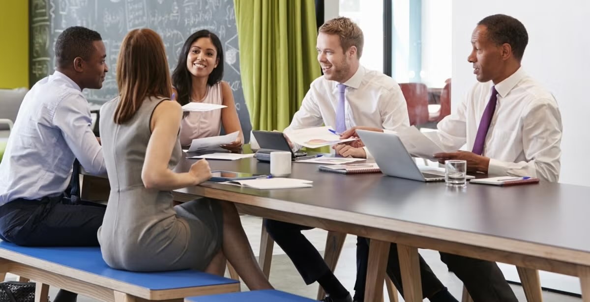A group of five professionals collaborating around a modern conference table, engaged in discussions with laptops and documents.