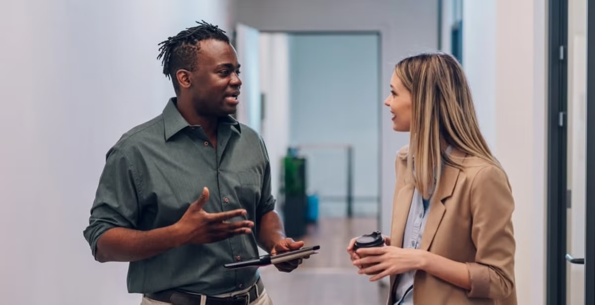 Two colleagues having a casual conversation in a hallway, one holding a tablet and the other a coffee cup.