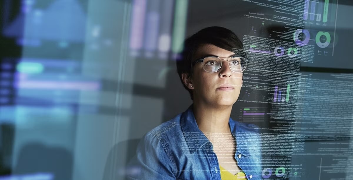 A woman with short hair and glasses, focused on data displayed on her computer screen in a dark room, analyzing complex visual data.
