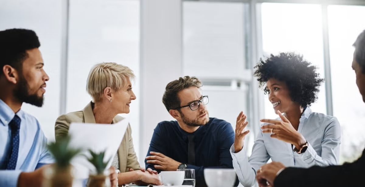 A diverse group of strategic communication professionals engaged in a discussion around a conference table, exchanging ideas with documents and laptops.