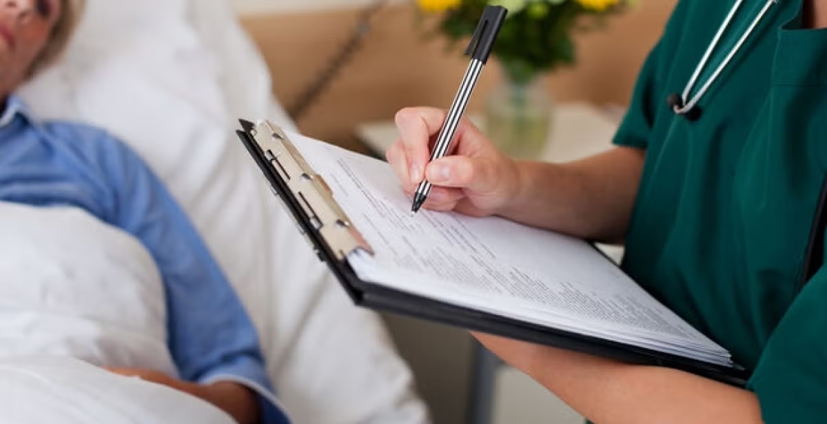 A healthcare professional writing on a clipboard while attending to a patient in a hospital bed.