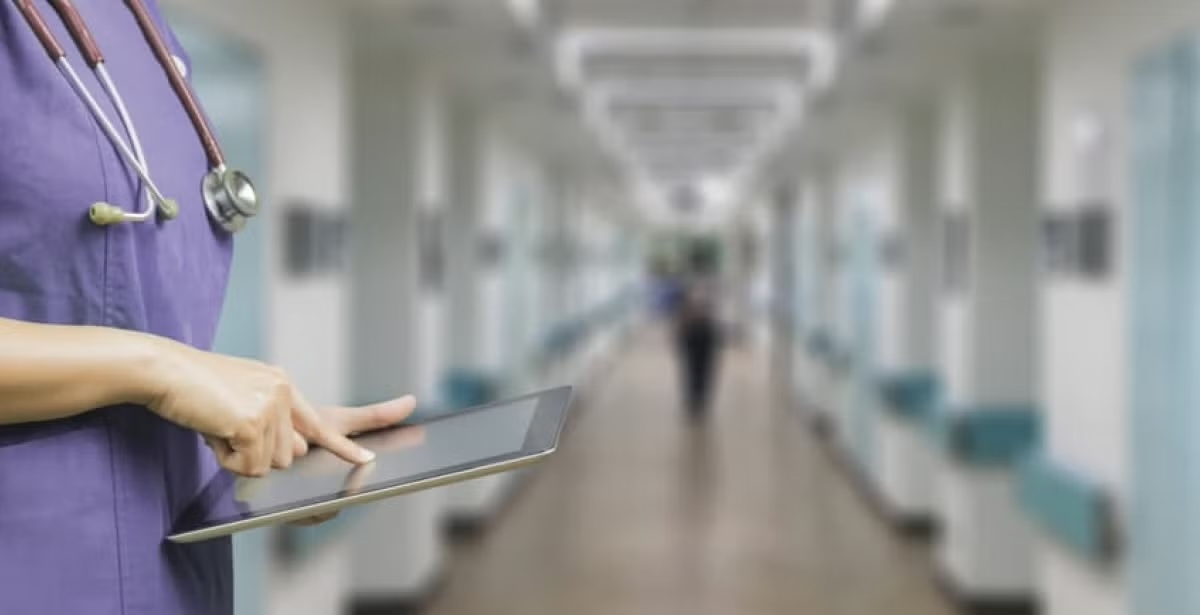 A healthcare professional in scrubs using a tablet in a hospital corridor.