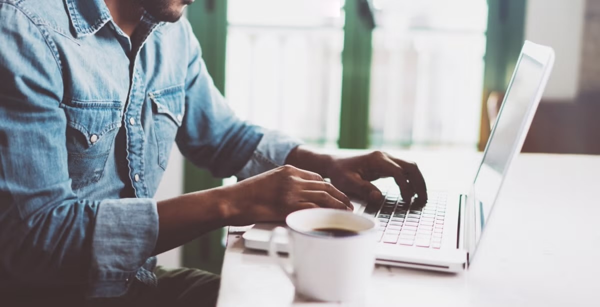 A man in a denim shirt typing on a laptop at a table with a cup of coffee nearby.