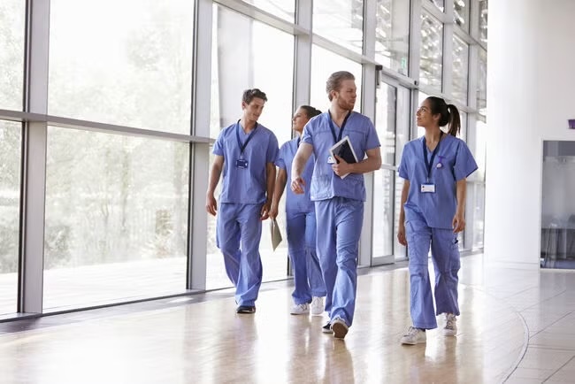 A group of medical professionals in scrubs walks through a bright hospital hallway, discussing patient care.