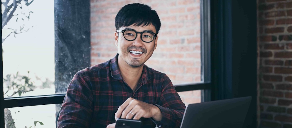 A smiling man with glasses sitting at a table with a laptop, holding a phone.