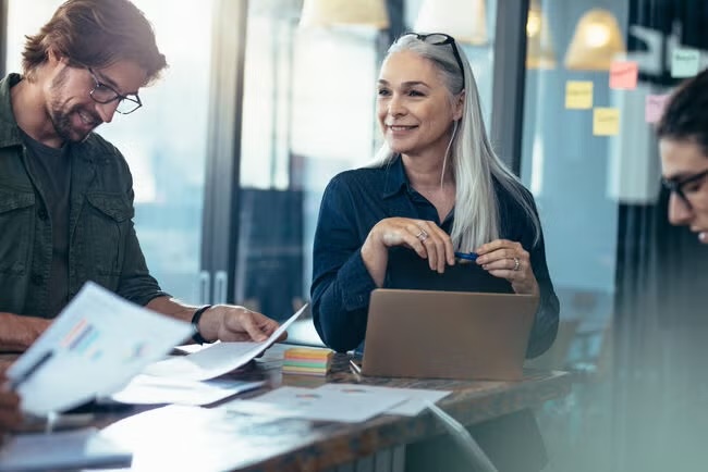 A senior woman leads a collaborative discussion with colleagues, sharing ideas and reviewing documents around a table.
