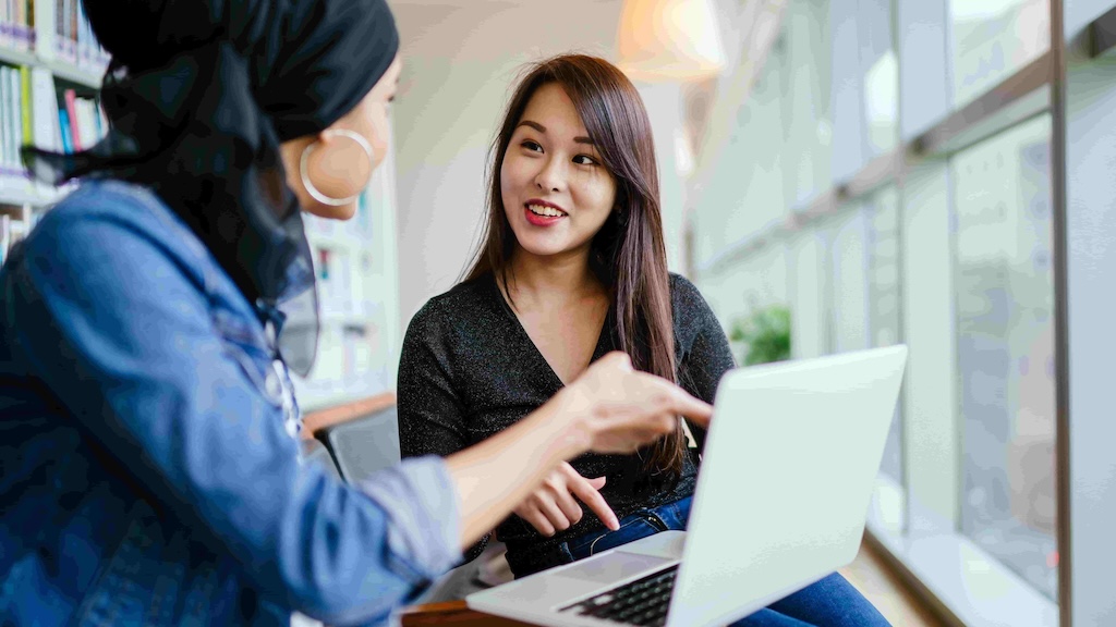 Two women in conversation while working on a laptop in a bright, modern space.