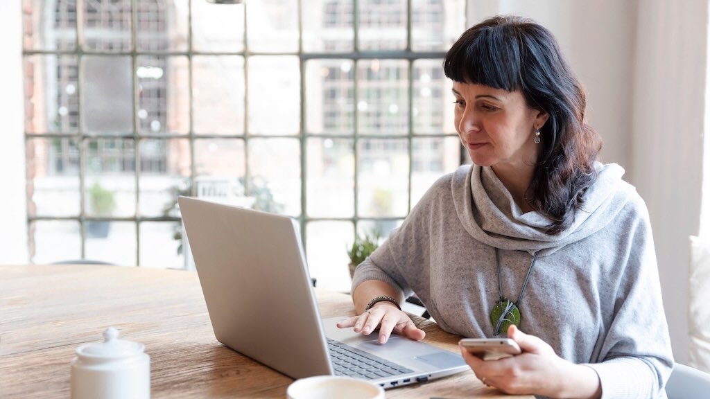 A woman working on a laptop at a table, holding a smartphone, in a relaxed indoor setting.