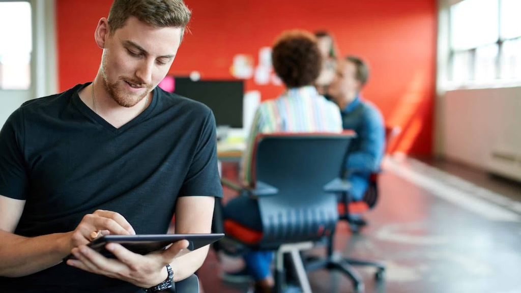 A young professional sitting in a modern office space, using a tablet.