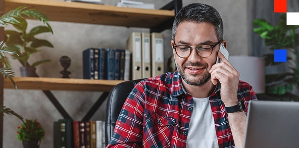 A man wearing glasses is seated at a desk, engaged in a phone conversation.