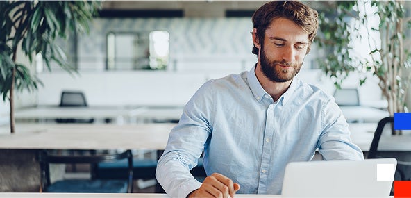 A man seated at a table, focused on his laptop, with a neutral background.