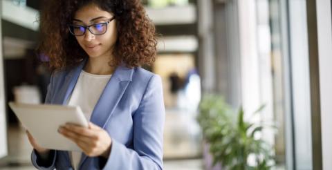 A businesswoman in a blue blazer, focusing on her tablet while standing in a bright office corridor, exemplifying modern workplace dynamics.
