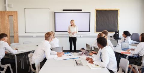 A teacher leading a classroom discussion with high school students, who are seated at desks with notebooks and laptops, attentively engaging in the session.