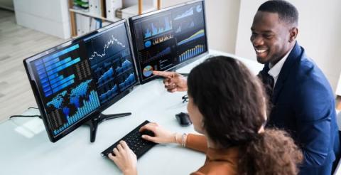 Two colleagues working together, analyzing graphs and data visualizations on dual computer monitors.