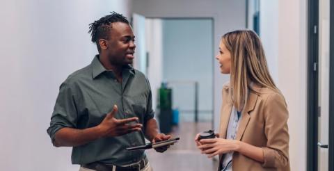 Two colleagues having a casual conversation in a hallway, one holding a tablet and the other a coffee cup.