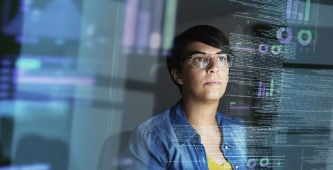 A woman with short hair and glasses, focused on data displayed on her computer screen in a dark room, analyzing complex visual data.