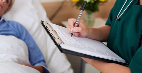 A healthcare professional writing on a clipboard while attending to a patient in a hospital bed.