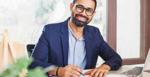 A smiling man wearing glasses and a blue jacket, working on papers and a laptop at his desk.