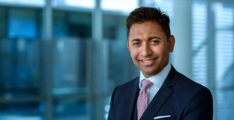 A well-dressed man with a confident smile standing in a modern office space, representing professionalism and leadership.