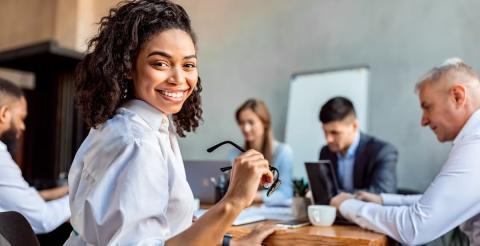 A smiling woman holding glasses during a business meeting with colleagues working in the background.