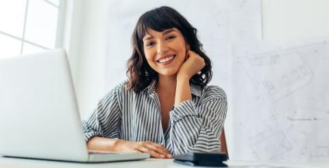 A woman smiling while working on a laptop in a bright office space, representing an urban designer at work.