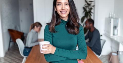 A smiling woman holding a cup while standing in a shared workspace, with two other people working in the background.
