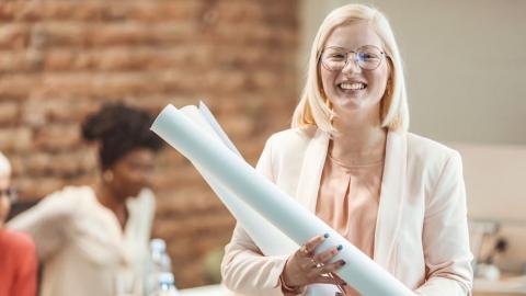 A cheerful woman holding blueprints, standing in an office setting, with colleagues in the background.