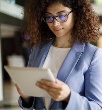 A businesswoman in a blue blazer, focusing on her tablet while standing in a bright office corridor, exemplifying modern workplace dynamics.