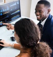 Two colleagues working together, analyzing graphs and data visualizations on dual computer monitors.