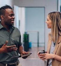 Two colleagues having a casual conversation in a hallway, one holding a tablet and the other a coffee cup.