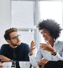 A diverse group of strategic communication professionals engaged in a discussion around a conference table, exchanging ideas with documents and laptops.