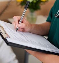 A healthcare professional writing on a clipboard while attending to a patient in a hospital bed.