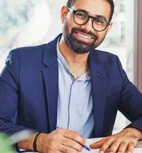 A smiling man wearing glasses and a blue jacket, working on papers and a laptop at his desk.