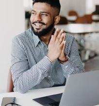 A man smiling and sitting at a desk with a laptop in a modern, open workspace.