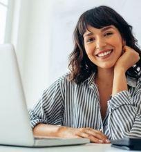 A woman smiling while working on a laptop in a bright office space, representing an urban designer at work.