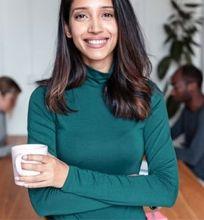 A smiling woman holding a cup while standing in a shared workspace, with two other people working in the background.