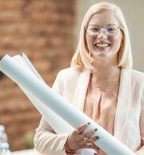 A cheerful woman holding blueprints, standing in an office setting, with colleagues in the background.