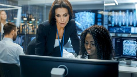 Female Team Leader Consults Young Computer Engineer. They Work in a Crowded Office on a Neural Network/ Artificial Intelligence Project.
