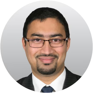 An Indian man wearing a formal suit and tie, standing confidently with a neutral expression.
