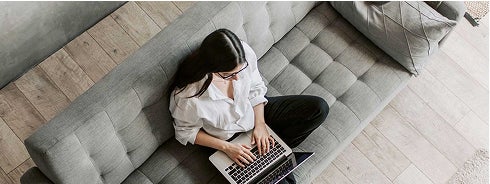A woman sitting comfortably on a couch, focused on her laptop, with a cozy living room setting in the background.